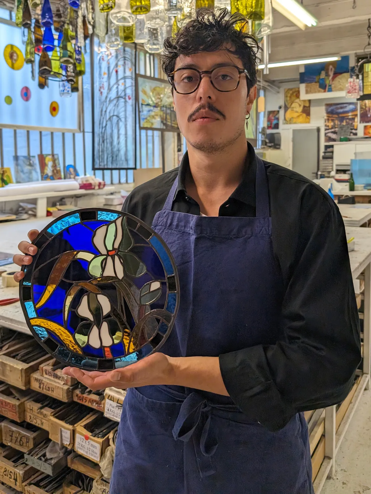 Andrés Pérez Flórez in the stained-glass workshop, holding a circular panel with a white iris motif on cobalt blue
