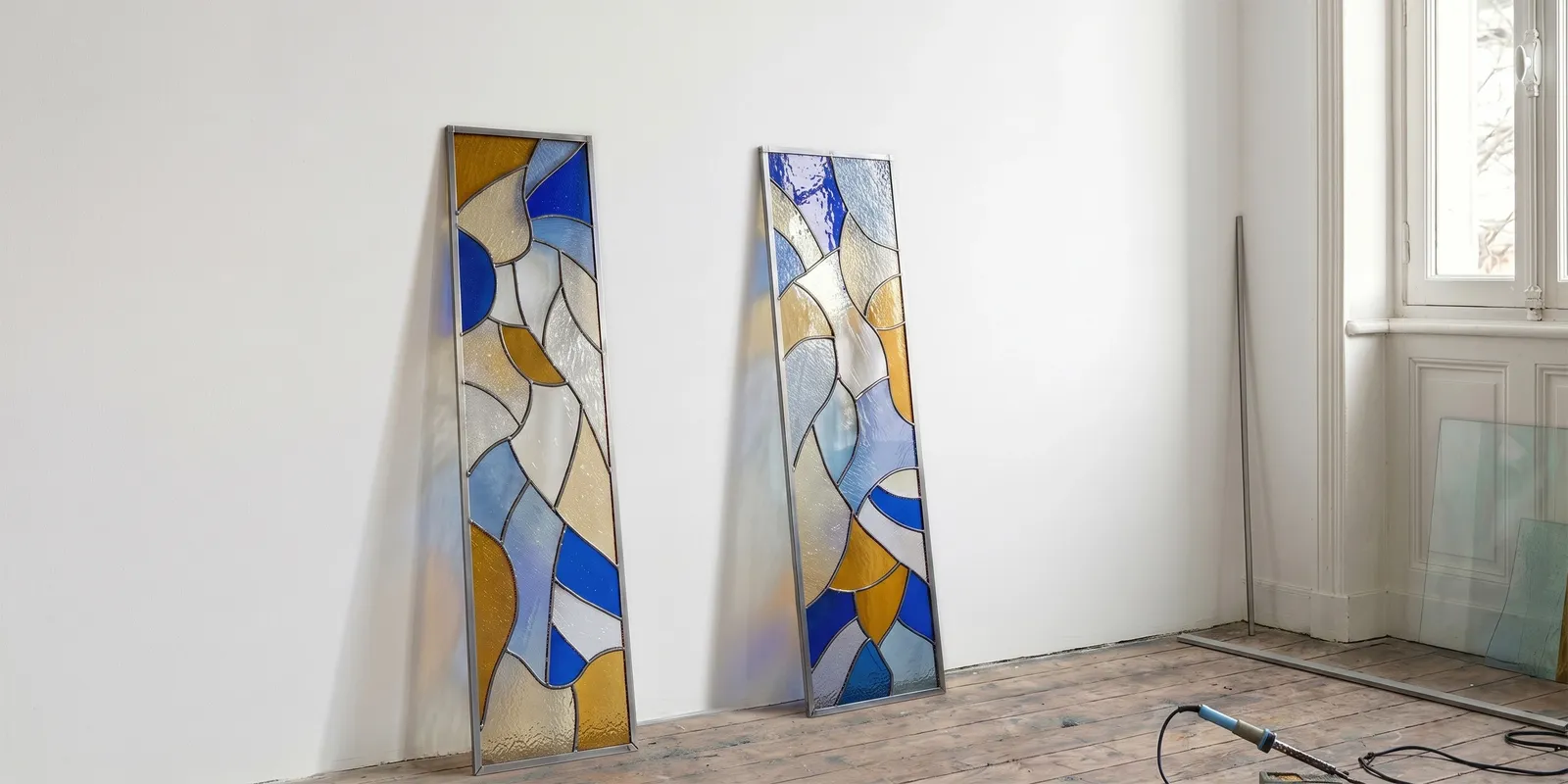 Fragments du ciel, two tall stained-glass panels in blue, amber and clear glass leaning against a white studio wall
